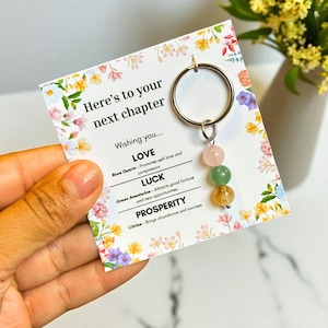 May include: A keychain with a silver ring and three gemstone beads: rose quartz, green aventurine, and citrine. The keychain hangs from a card with floral accents and the text "Here's to your next chapter". The card also lists the properties of each gemstone.