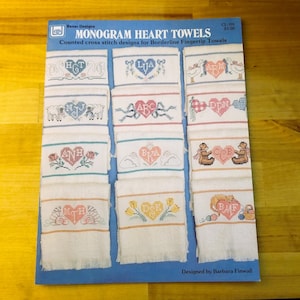 May include: A blue cover for a cross-stitch pattern book titled "MONOGRAM HEART TOWELS." The cover displays several white fingertip towels with embroidered heart designs, each featuring initials and decorative elements like swans, sheep, and flowers.