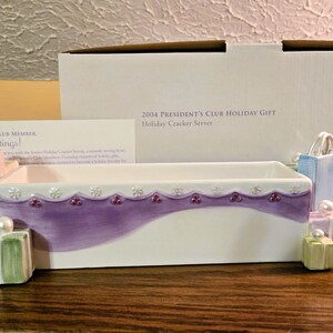 May include: A rectangular white ceramic serving dish with a purple wavy painted accent, decorated with small sculpted gift boxes in pastel colors, pearl-like spheres, and snowflake details. The dish is accompanied by its original white cardboard box.