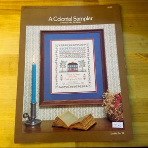 May include: A brown and white booklet with the title "A Colonial Sampler" and a picture of a cross-stitch sampler with the alphabet, numbers, and a small house with the text "Peace to those who enter, God's grace to those who depart."
