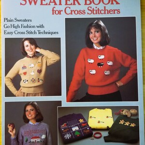 May include: A book titled "The Sweater Book for Cross Stitchers" by Anne Van Wagner Young. The cover features photos of women wearing sweaters with cross-stitch designs, including a red sweater with sheep, a beige sweater with a house and other small designs, and a blue sweater with an alphabet design.