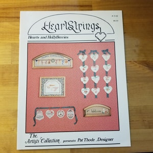 May include: A HeartStrings cross stitch pattern booklet featuring a variety of heart-shaped designs with a red and white color scheme. The booklet includes a welcome sign, a Merry Christmas sign, and a variety of heart ornaments. The booklet is titled "Hearts and HollyBerries" and is designed by Pat Thode.
