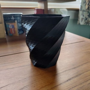 Può includere: Una tazza o un vaso nero stampato in 3D con un motivo geometrico. La tazza ha un design a spirale contorto.