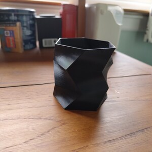 Può includere: Un contenitore nero di forma geometrica con una superficie testurizzata. Il contenitore è realizzato in materiale plastico rigido e ha una forma esagonale.