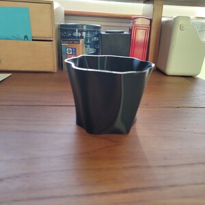 May include: A black, wavy, plastic planter pot on a wooden surface.