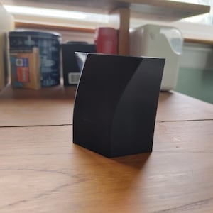 Può includere: Un oggetto rettangolare nero stampato in 3D con un lato curvo. L'oggetto è su una superficie di legno.