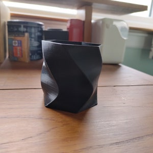 May include: A black, geometric, 3D printed planter with a twisted design. The planter is sitting on a wooden surface.
