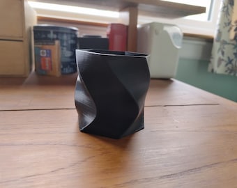 May include: A black, geometric, 3D printed planter with a twisted design. The planter is sitting on a wooden surface.