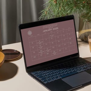 May include: A laptop computer with a calendar on the screen. The calendar is for January 2025 and has a pink background. The laptop is on a white table with a bowl of nuts and a glass of tea.