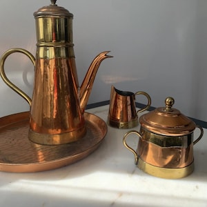 May include: A copper coffee pot set with brass accents. The set includes a large coffee pot on a tray, a creamer, and a sugar bowl. The coffee pot has a long spout and a handle. The creamer and sugar bowl have small handles.