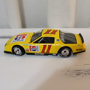 May include: Yellow die-cast model car with a black spoiler, Pepsi logo, and the number 11 on the side. The car is on a white surface.