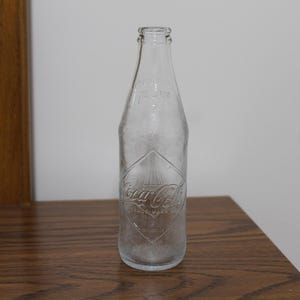 May include: A clear glass Coca-Cola bottle with a diamond-shaped logo embossed on the front.