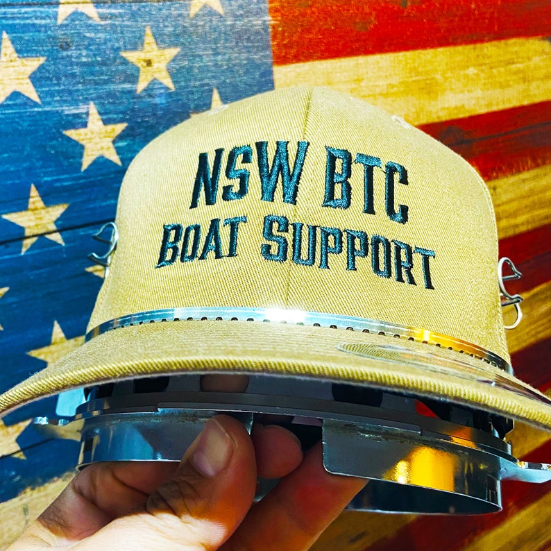 May include: A tan baseball cap with a black embroidered logo that reads "NSW BTC BOAT SUPPORT". The cap has a silver band around the crown and a silver buckle on the back.