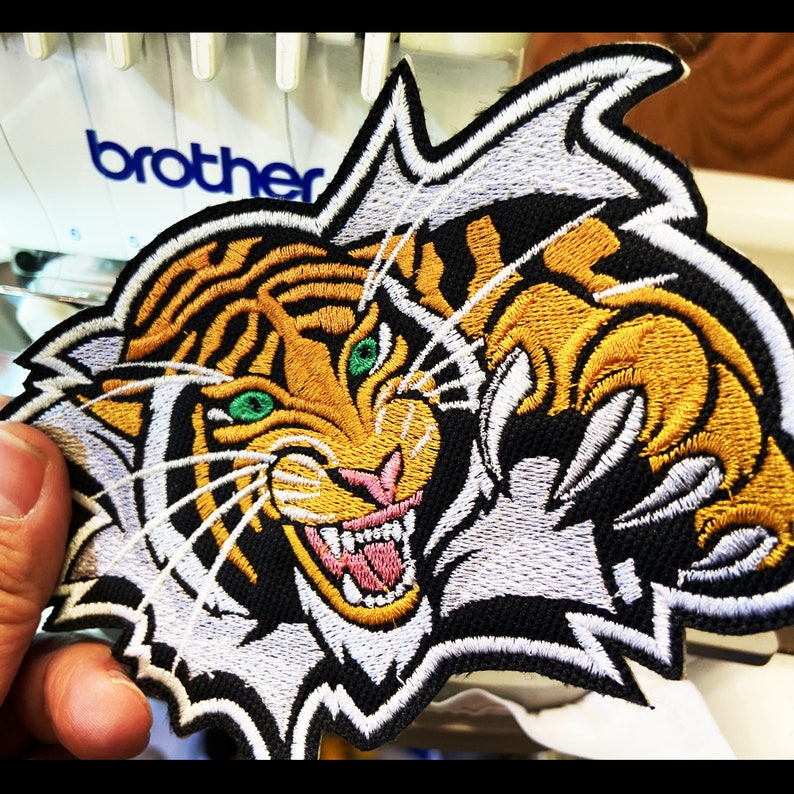 May include: Embroidered iron-on patch featuring a tiger's face with green eyes and a snarling mouth. The tiger is breaking through a white background with its claws.