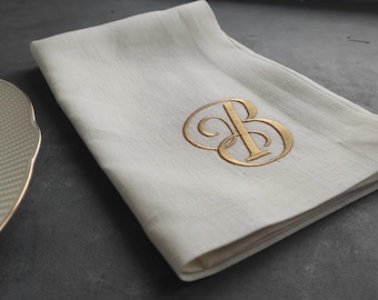 May include: A folded white fabric napkin featuring an embroidered gold monogram of the letters B and O intertwined in a decorative script font. The napkin is positioned next to a white plate with a textured rim and a gold-colored edge.