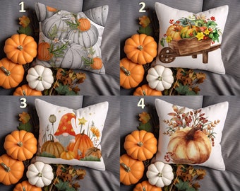 May include: Four decorative throw pillows with fall-themed designs. Pillows feature pumpkin and autumn leaf illustrations. Surrounding the pillows are orange and white pumpkins, and fall foliage. The pillows are on a gray surface.