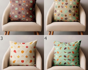 Può includere: Quattro cuscini decorativi con motivi autunnali. Ogni cuscino presenta un design diverso di foglie, ghiande e bacche in vari colori, tra cui marrone, arancione e verde, su uno sfondo neutro. I cuscini sono posizionati su una sedia beige.