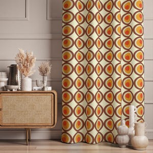 Op de afbeelding: Crèmekleurig gordijn met een herhalend patroon van bruine, oranje en gele ovale vormen. Het retro-ontwerp voegt kleur en visuele interesse toe aan elke kamer. Het gordijn is gemaakt van een zachte, vloeiende stof.