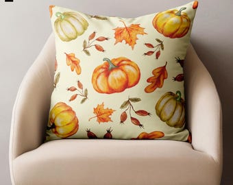 May include: Square decorative pillow with a cream background featuring a watercolor pattern of pumpkins, orange and yellow fall leaves, and sprigs of berries. The pillow is placed on a beige chair.