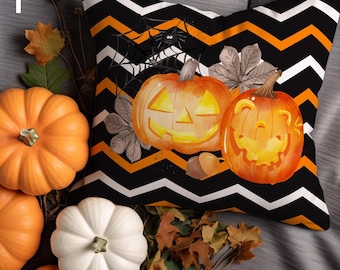 May include: A Halloween-themed decorative pillow with a black and orange chevron pattern. The pillow features two carved pumpkins, a spider, and fall foliage. Several orange and white pumpkins and leaves surround the pillow.