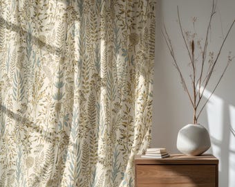 Puede incluir: Cortina color crema con un estampado botánico con hojas y tallos verdes y azules. La cortina cuelga junto a una mesita de noche de madera con un jarrón y ramas decorativas. El diseño de la cortina se repite.