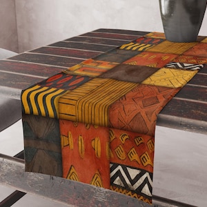 Op de afbeelding: Decoratieve tafelloper met een patchwork ontwerp. De loper heeft een verscheidenheid aan geometrische patronen in tinten oranje, geel, bruin en zwart. De loper wordt gepresenteerd op een donkere houten tafel.