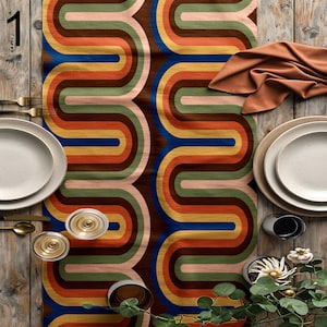 Può includere: Un runner da tavola con un motivo geometrico ondulato ripetuto nei toni del marrone, arancione, giallo, blu, verde e rosa chiaro. Il runner è presentato con piatti bianchi, posate dorate, bicchieri e un tovagliolo arancione piegato.