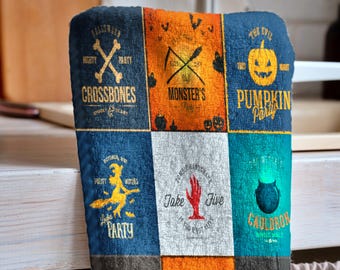 May include: A Halloween-themed towel with various square panels featuring spooky graphics and text. Designs include crossbones, pumpkins, witches, cauldrons, ghosts, and spiderwebs. The color palette includes navy blue, orange, gray, and teal.