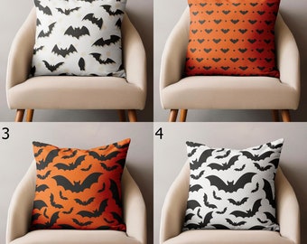 May include: Four decorative throw pillows with Halloween-themed bat patterns. The pillows feature designs in black and orange, and black and white. The pillows are square and placed on beige chairs.
