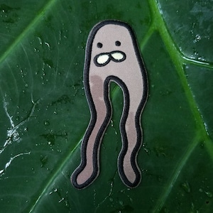 May include: A brown felt patch with a cartoon character with a smiling face and two legs. The character is outlined in black.