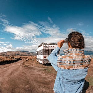 Puede incluir: Una persona con una chaqueta vaquera azul con cuello estampado, mirando el paisaje. Una autocaravana blanca con una escalera está estacionada en un camino de tierra. El cielo es azul con nubes blancas y montañas al fondo.