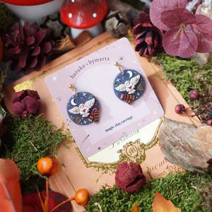 Pode incluir: Brincos redondos azuis com um desenho de coruja branca, uma lua crescente e uma carta. Os brincos estão em um cartão rosa com o texto "haruko.bymarta" e "magic clay earrings". O fundo inclui folhagem de outono e um cogumelo.