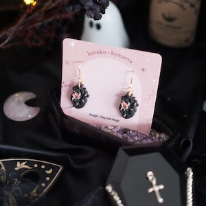 Könnte beinhalten: Ein Paar schwarze herzförmige Ohrringe mit rosa floralen Akzenten, präsentiert auf einer rosa Karte mit dem Text "haruko - bymarta" und "magic clay earrings".