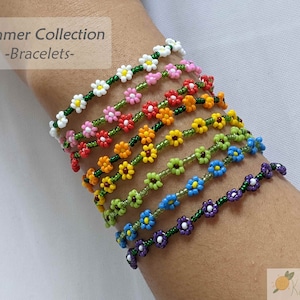 May include: A stack of colorful beaded bracelets with flower designs. Each bracelet features a different color of flower, including white, pink, orange, yellow, green, blue, and purple. The bracelets are all strung on green thread.