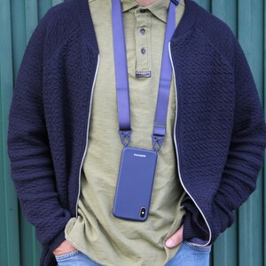 May include: A navy blue phone case with a matching strap, hanging around the neck. The phone case has the brand name "HANUPP" printed on it. The person is wearing a navy blue jacket, a green shirt, and blue jeans.