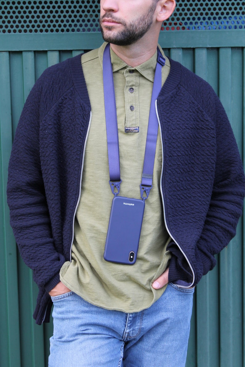 May include: A navy blue phone case with a matching strap, hanging around the neck. The phone case has the brand name "HANUPP" printed on it. The person is wearing a navy blue jacket, a green shirt, and blue jeans.