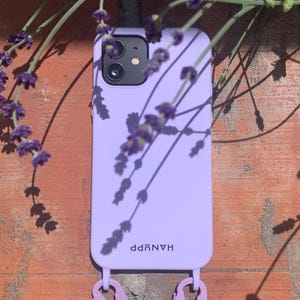 May include: A lavender phone case with the word "HANUPP" printed on it. The case has two lavender-colored clips attached to it. The phone case is resting against a brick wall, with lavender flowers in the background.