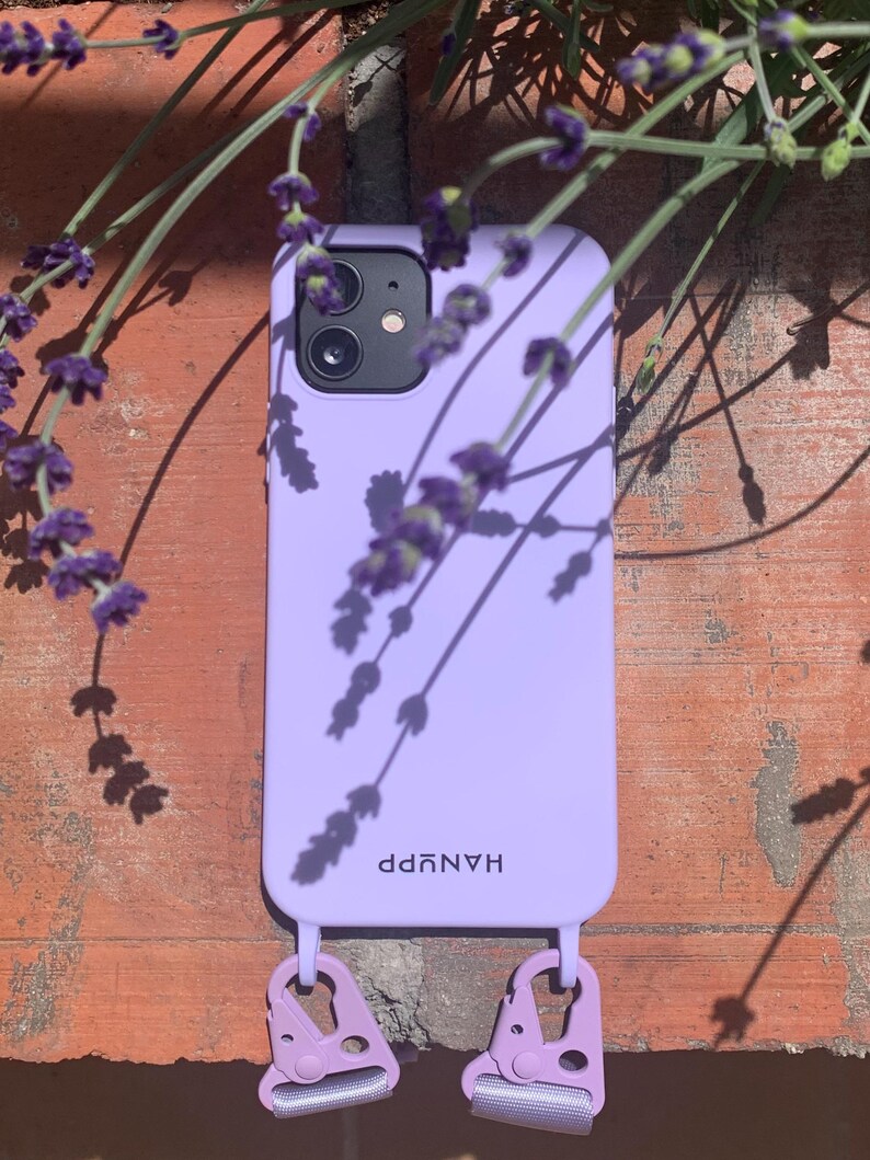 May include: A lavender phone case with the word "HANUPP" printed on it. The case has two lavender-colored clips attached to it. The phone case is resting against a brick wall, with lavender flowers in the background.
