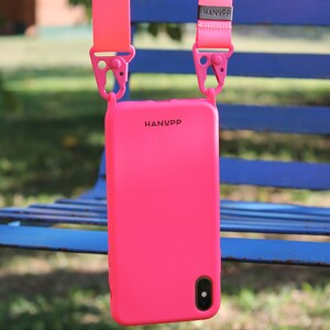 May include: Bright pink phone case with a matching strap, hanging against a blue bench. The case has the brand name "HANYPP" printed on it. The strap is attached with pink clips.