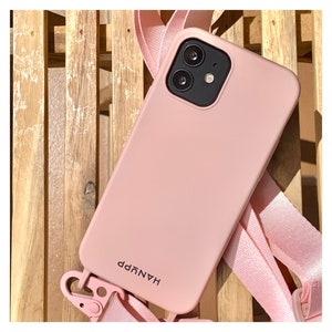 May include: A light pink phone case with a matching strap, resting on a wooden surface. The case has a dual-lens camera and the word "HANYPP" printed on it. The strap is attached to the case with a pink clasp.