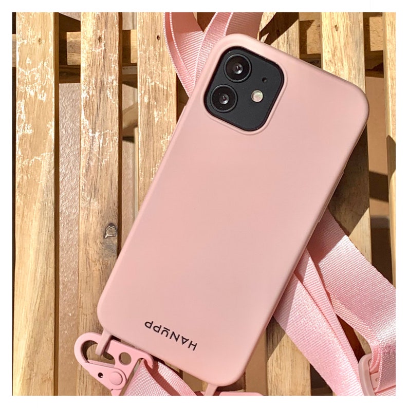 May include: A light pink phone case with a matching strap, resting on a wooden surface. The case has a dual-lens camera and the word "HANYPP" printed on it. The strap is attached to the case with a pink clasp.