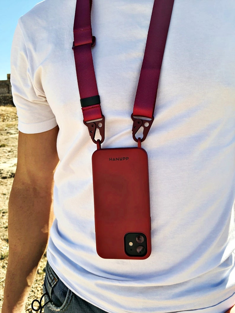 May include: A red phone case with the word "HANYPP" printed on it, attached to a burgundy lanyard. The lanyard has metal clips and is worn around the neck. The phone case has a dual-lens camera.