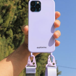 May include: A light purple phone case with a matching strap. The case has the word "HANUPP" printed on it. The strap is attached with lavender-colored clips. The background is a bright blue sky and green foliage.