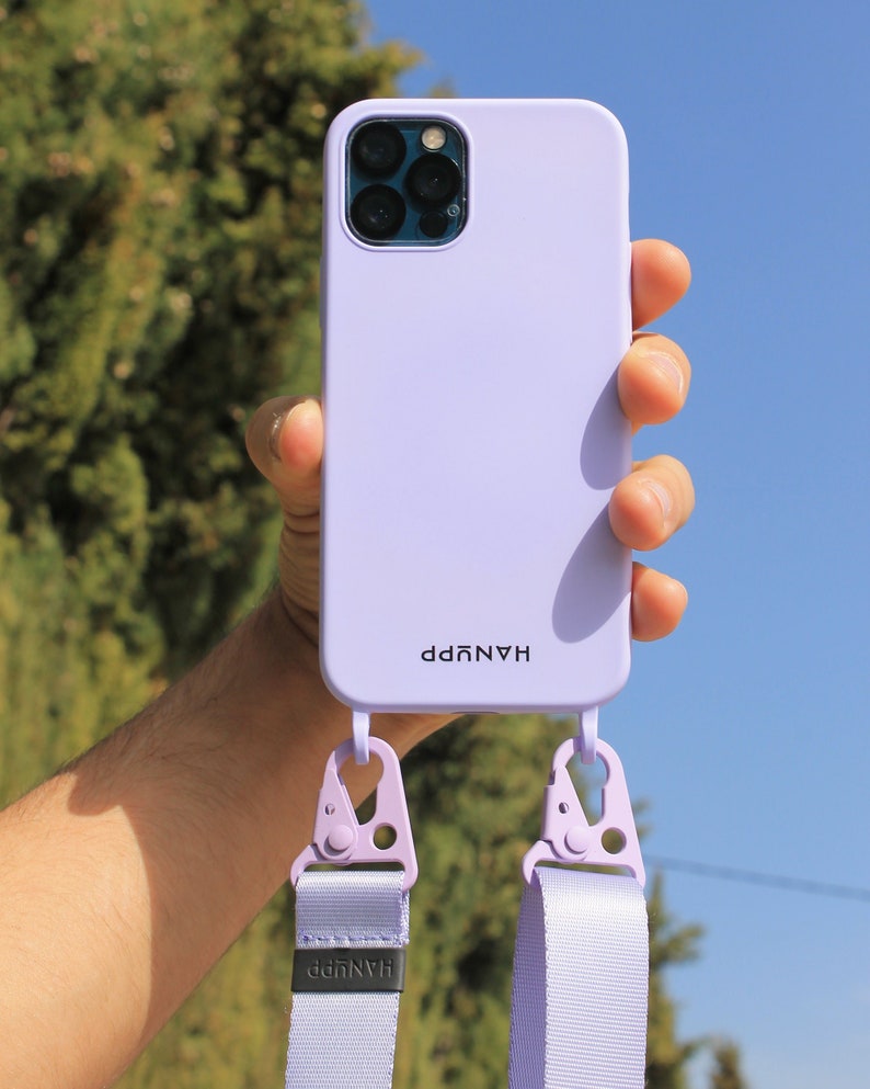 May include: A light purple phone case with a matching strap. The case has the word "HANUPP" printed on it. The strap is attached with lavender-colored clips. The background is a bright blue sky and green foliage.