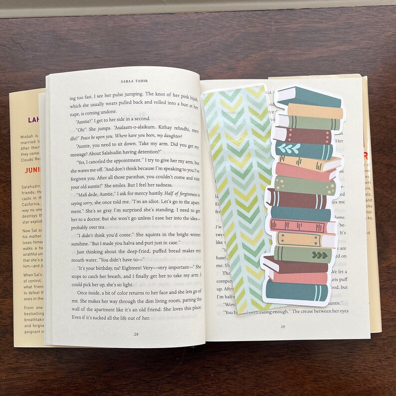 Peut inclure: Un livre ouvert avec un marque-page repr&eacute;sentant une pile de livres color&eacute;s. Le marque-page est un autocollant avec un motif chevron vert et blanc. Les pages du livre pr&eacute;sentent du texte d'un roman sur une jeune femme et sa tante.