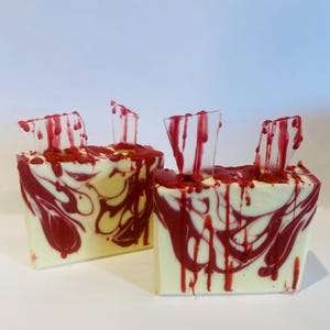 May include: Two bars of handmade soap with a white base and red swirls. The soap is topped with clear glass shards and red dripping wax.