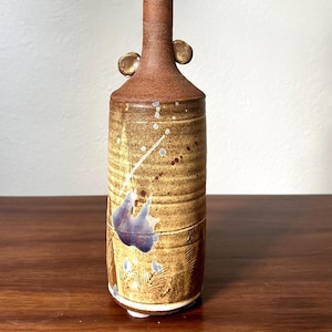 May include: A handcrafted ceramic vase with a tall, slender neck and a cylindrical body. The vase features a brown and tan color scheme with blue and white accents. It has two small, rounded handles near the neck.
