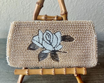 May include: A vintage beaded handbag with a bamboo handle and stand. The bag is covered in small, cream-colored beads and features a white and brown beaded rose design on the front. The bamboo handle and stand add a natural touch to the elegant design.