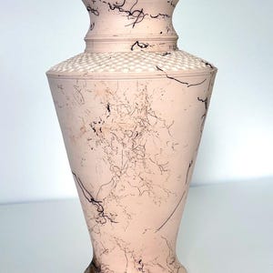 May include: A tall, pale pink vase with a flared base and a wide, textured rim. The vase features a series of black, crack-like lines across its surface. The middle section has a dotted pattern. The vase is a decorative item.