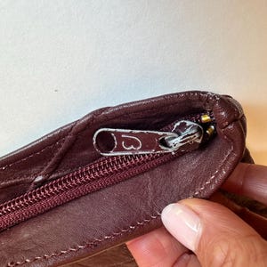 May include: Close-up of a burgundy leather bag with a silver zipper. The zipper pull has a heart design. The bag's leather has a quilted pattern and is stitched with burgundy thread. The bag is partially open, revealing the interior.
